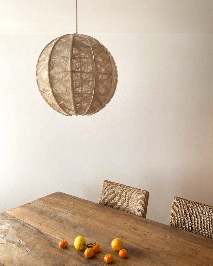 eine runde Hängeleuchte aus Birkenholz umwebt mit beigem Leinengarn hängt über einem Holztisch mit Zitrusfrüchten